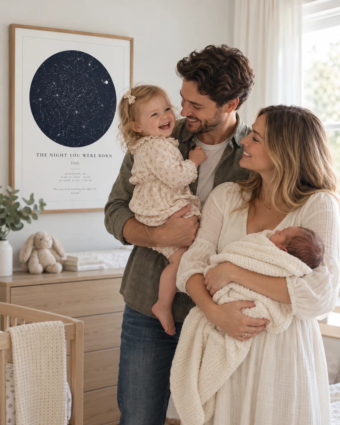 New parents holding their newborn and toddler beside a framed star map titled The Night You Were Born