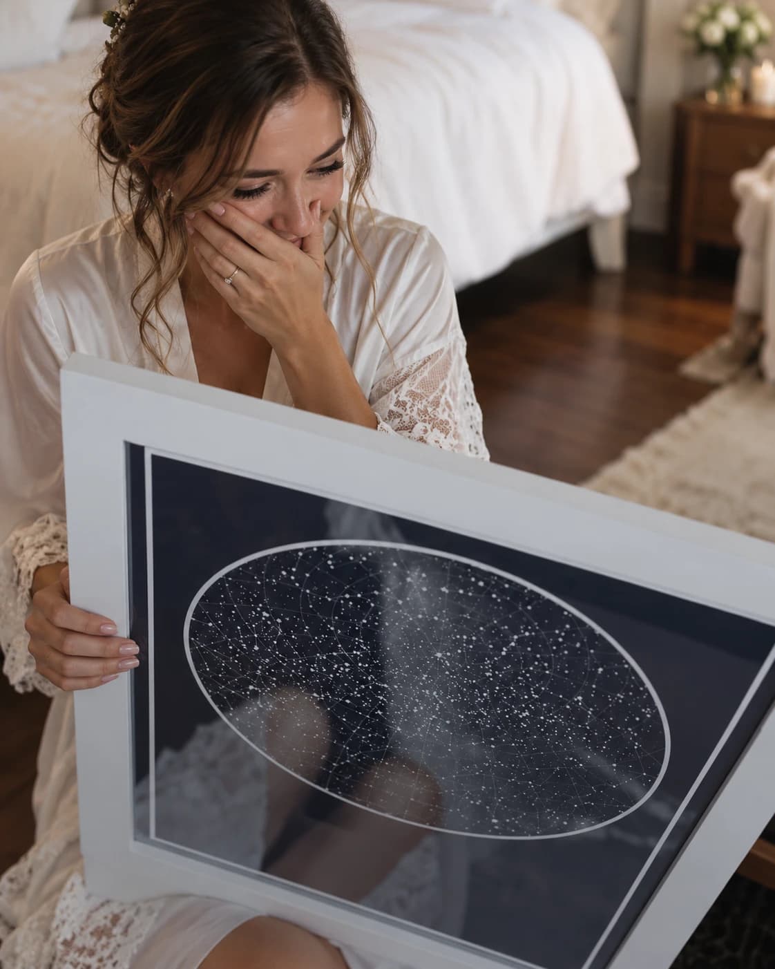 A bride in her robe, hand to her face, looking down at a framed navy star map she has just been given